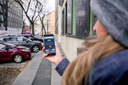 Frau mit einem Handy in der Hand geht durch eine Straße. Es ist Winter. Grauer Himmel und nackte Bäume sind zu sehen.