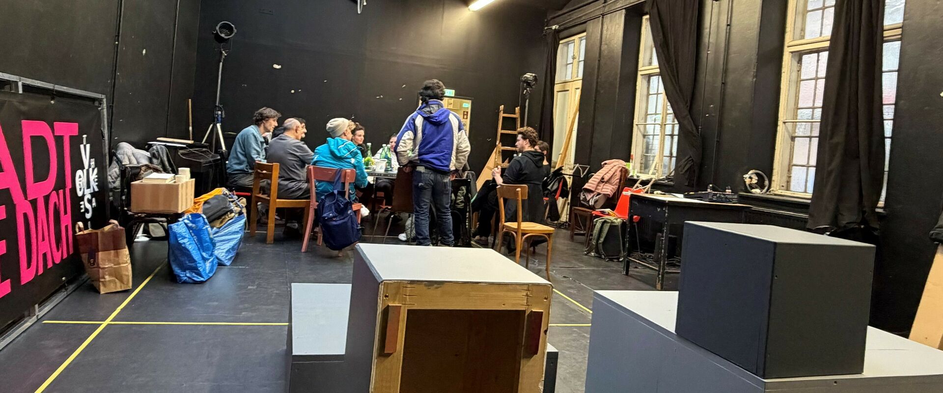 Menschen um einen Tisch im schwarz gestrichenen Proberaum. An der Wand lehnt der Titel des Stückes "Stadt ohne Dach". 