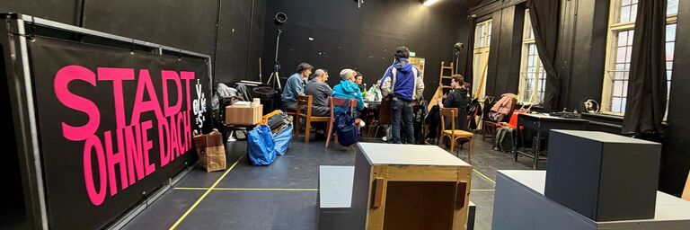 Menschen um einen Tisch im schwarz gestrichenen Proberaum. An der Wand lehnt der Titel des Stückes "Stadt ohne Dach". 