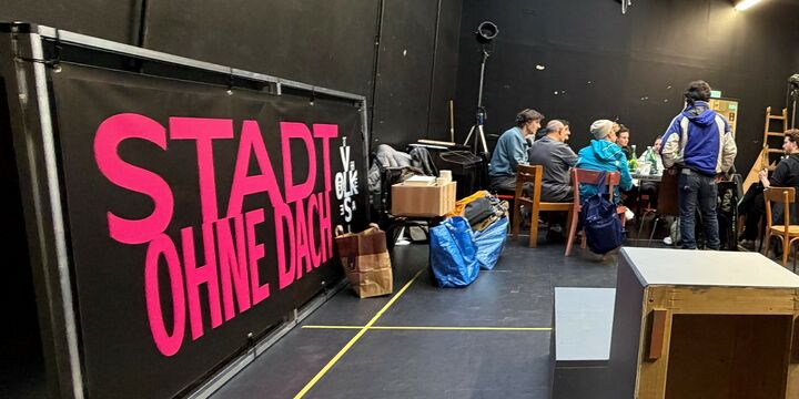 Menschen um einen Tisch im schwarz gestrichenen Proberaum. An der Wand lehnt der Titel des Stückes "Stadt ohne Dach". 