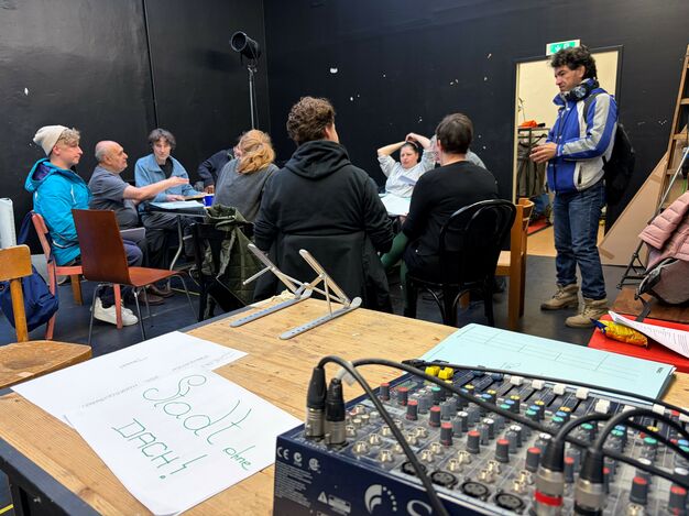 Menschen sitzen um einen Tisch in einem schwarz gestrichenen Raum. Eine Person in Jacke kommt herein. Im Vordergrund ist ein Mischpult zu sehen.