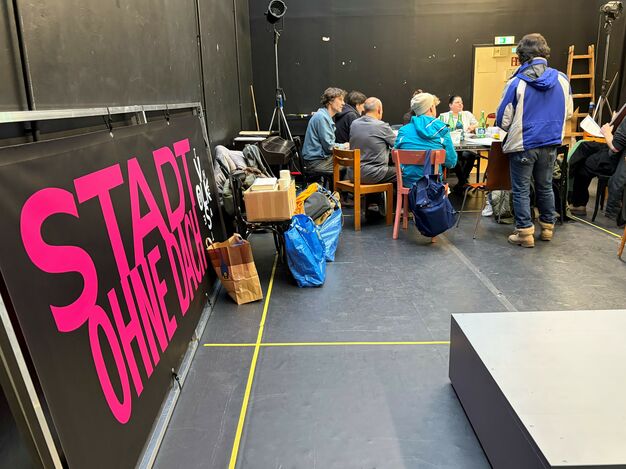 Menschen an einem großen Tisch in einem schwarzen Proberaum. An der Wand lehnt der Schriftzug des Stückes. 