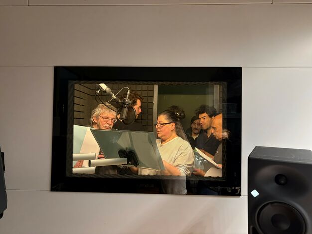 Das Fenster in den Aufnahmeraum des Tonstudios, in dem sieben Personen, Mikrofone und Textbögen zu sehen sind,