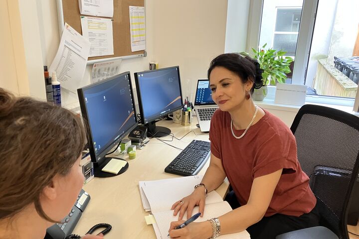 Im Obdach Dreyhausenstraße finden geflüchtete Menschen Beratung im Alltag, zu Gesundheit und bei Behördenangelegenheiten. (Bild: FSW) Beratungssituation: Eine Frau sitzt mit dem Rücken zur Kamer und lässt sich zu einer Frage beraten. Die Beraterin sitzt am Schreibtisch. Computer und Unterlagen sind zu sehen.