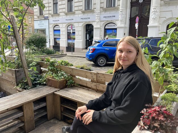 Diana Schmidt zwischen den Pflanzen und Sträuchern der Grätzeloase vor dem Tageszentrum Obdach aXXept. (Bild: FSW) Diana Schmidt sitzt auf einer Bank, die Teil einer Holzkonstruktion aus Sitzgelegenheiten und Blumenkästen ist. Im Hintergrund sind Stadtgebäude und geparkte Autos zu sehen.