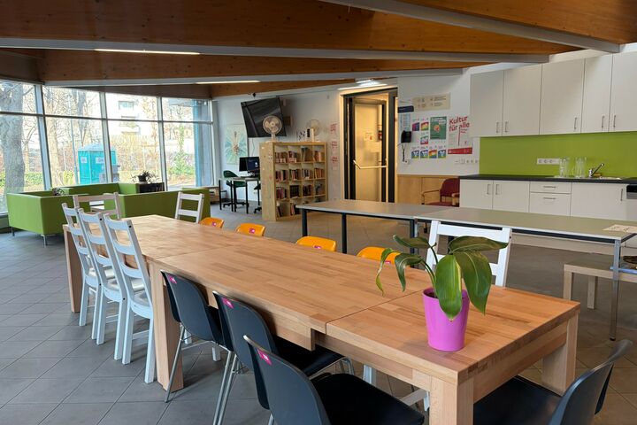 Eine große Tafel mit unterschiedlichen Sesseln und einer Topfpflanze. Im Hintergrund ist eine Küche, eine Sitzlandschaft, offene Bücherschränke und Tische mit Computern zu sehen,