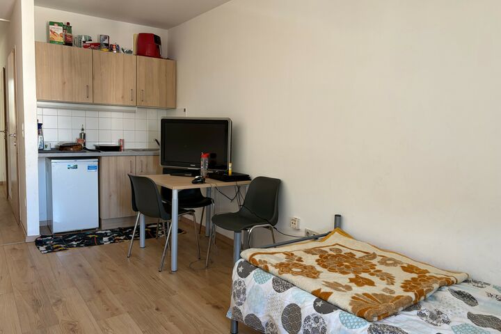 Eine Kochnische, davor ein Tisch mit einem Fernsteher und ein Bett. 